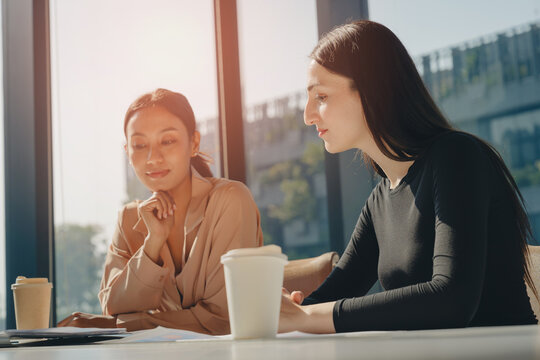 Two young women working together in bright office with large windows, sharing ideas and enjoying coffee, creating focused and collaborative atmosphere