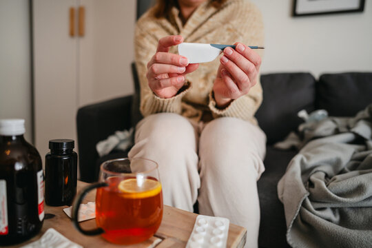 Person with flu and fever holding thermometer on sofa with medicine