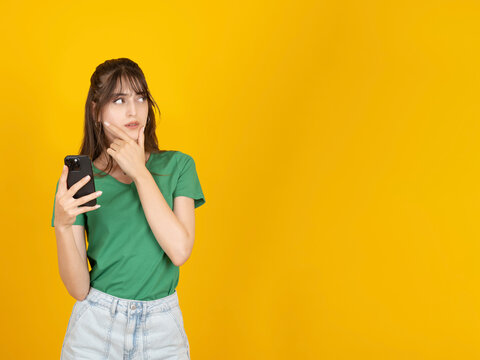 Young woman holding smartphone while touching chin thoughtfully, wearing green shirt and denim shorts, standing against bright yellow studio background with large copy space. People communication.