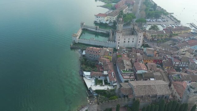 Drone flyaway from Scaliger Castle in Sirmione, Italy, revealing turquoise waters of Lake Garda, colorful houses, and historic walls on a winter day.