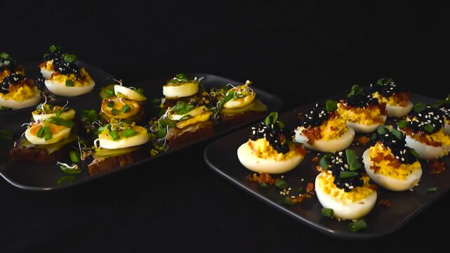 Studio shot of traditional Estonian appetizers including deviled eggs topped with caviar and bacon and rye bread canapes with sprat fish egg slices pickles and microgreens on dark background.