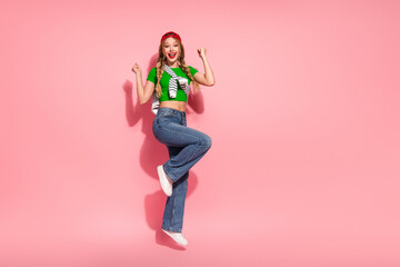 Joyful young woman jumps in pink studio wearing green top jeans cap smiling bright