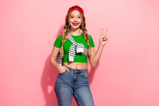 Bright stylish young woman with braids in a green crop top and jeans posing with peace sign against pink background