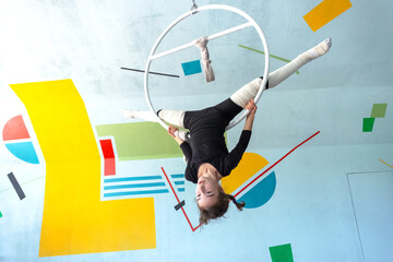 Young acrobat girl stretching upside down on hoop, circus studio, elastic protection, performing acrobatic trick, safe environment.