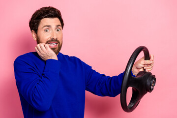 Young man in blue sweater holds steering wheel with surprised smile against pink background for...