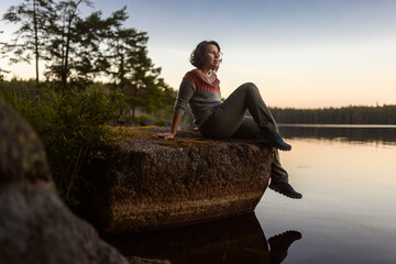A serene woman sits on a mossy rock by a calm lake at sunset, wearing a traditional Nordic knit...