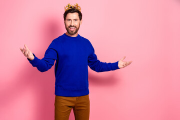 Confident young man wearing blue sweater with crown smiles and poses against pink background
