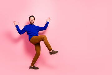 Naklejka premium Happy young man in blue sweater and brown pants leaps with arms raised against pink background expressing energy joy and a casual lifestyle for fashion advertising