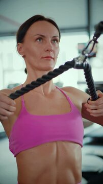 Vertical. A woman in a pink sports bra performs a cable row exercise in a bright, modern gym. Focused woman pulls on gym cables for a back workout in a fitness studio. Athletic woman training 