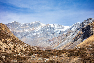 Obraz premium Wide panoramic view of the Himalayan mountain range featuring towering snow-capped peaks rising above a rugged alpine valley. The dramatic landscape showcases steep rocky slopes