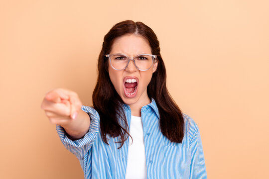 Angry young woman yelling and pointing finger directly at camera in confrontational portrait with glasses and striped shirt on peach background