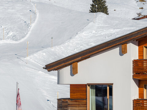 Close up winter view of a modern chalet style hotel in Lech Oberlech, Austria, with circular mountain logo emblem, wooden balconies, piste poles, and a lone evergreen.