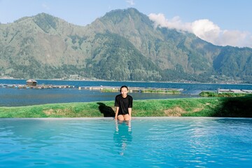 Fototapeta premium Woman in black swimsuit standing in pool with mountain and lake background