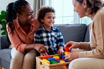 Black woman and Caucasian woman interacting with smiling child playing with colorful plastic building blocks on living room sofa, both adults engaging with young boy during playtime