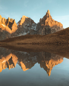 View of jagged peaks ablaze with the warm glow of the setting sun, mirrored in the glassy surface of a tranquil lake, Passo Rolle, Trentino-Alto Adige, Italy.