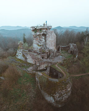 Aerial view of weathered stone ruins of a medieval castle atop a hill, a testament to time's passage, against a backdrop of misty, muted greens, Wingen, Grand Est, France.