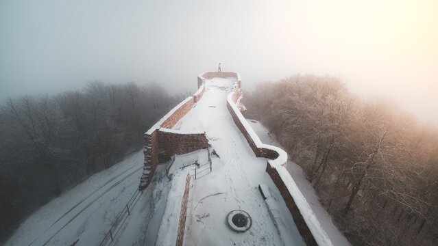 View of snow-covered ancient fortress walls vanishing into the dense winter fog, contrasting with the stark trees, Chateau du Wegelnburg, Germany.