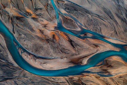 Aerial view of glacial rivers carve intricate paths through volcanic sands, a tapestry of turquoise against the stark grey and ochre landscape, Iceland.