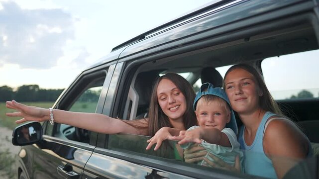 Happy family waves from car window during road trip vacation. Mother with children smile in vehicle. Family enjoys summer travel. Kids wave from car on journey. Road trip with happy family together.