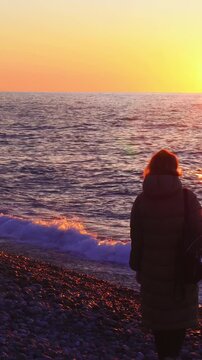 Woman walking along rocky beach at sunset, ocean waves gently lapping shore