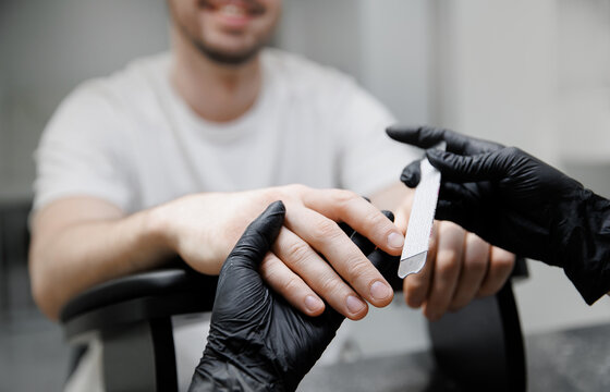 Manicurist woman using nail file and filing young adult man nails. Manicure, pedicure beauty salon concept. Care about fingernails of men