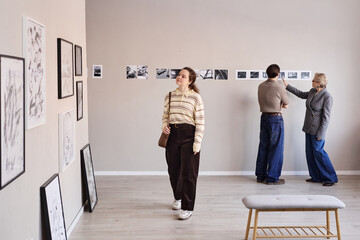 Fototapeta premium Young adult Caucasian woman walking through art gallery, middle aged Caucasian man and senior woman examining black and white photographs displayed on wall