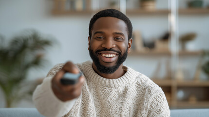 Fototapeta premium African american man holding television remote control and smiling. Relaxed guy watching show at home. Leisure activity in living room, entertainment and media consumption concept.