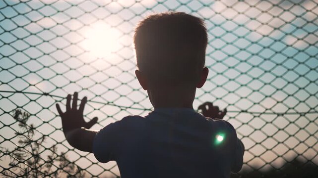 Boy reaching hands through chainlink fence toward sunlight outdoors. Child raises arms against fence barrier. Silhouette boy reaches through chainlink. Child lifts hands at wire fence with sunlight.