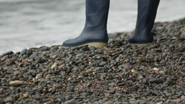 Boots, rubber, pebbles show closeup of person standing on rocky beach near crashing ocean waves, durable footwear