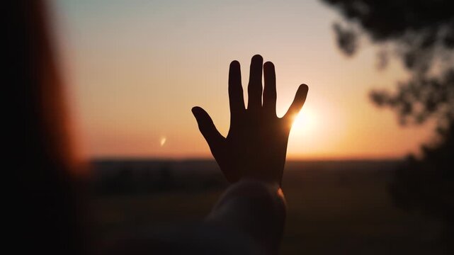 Woman raises hand toward sunset creating silhouette at golden horizon. Person reaches palm against sun at dusk. Female hand blocks sunlight. Woman shows gesture at sunset. Silhouette against sun.