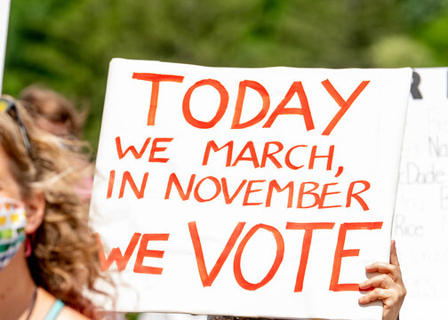 A protestor holds a bright red handmade sign reading "Today We March, In November We Vote," connecting street activism, civic engagement, and electoral action.