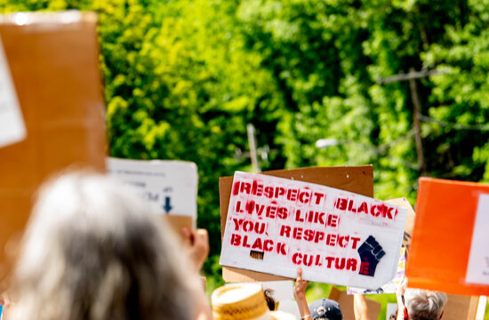 A handmade protest sign bearing the words "Respect Black lives like you respect Black culture" is raised above the heads of a crowd of anonymous marchers in a civil rights demonstration. 