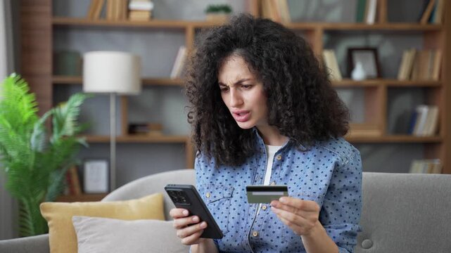 Shocked upset woman faced fraud checking credit card on mobile phone sitting in living room at home. Frustrated woman became a victim of deception, money had been stolen from her account. Close up