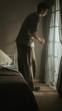 Vertical shot of apathetic Caucasian man sitting down on bed, drinking morning coffee from cup and looking out of window after sleepless night