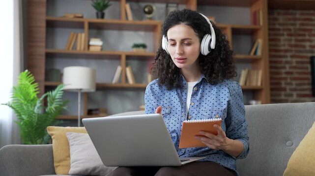 Woman in headphones watching video call training notes in notebook looking at laptop computer screen sitting on sofa in living room at home. Female listens to online learning, communicates remotely