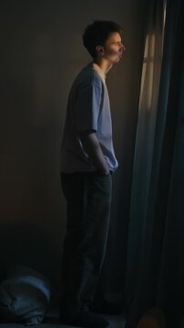 Vertical long shot of apathetic Caucasian man standing by window while spending sleepless night alone in bedroom