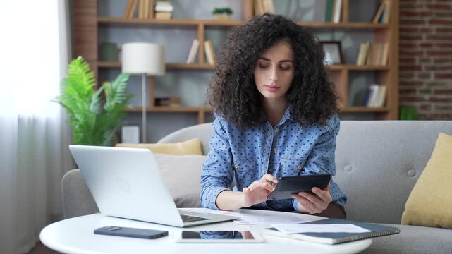 Young busy woman dealing with household finances sitting on sofa in living room at home office. Beautiful female doing paperwork, calculates on a calculator, writes down data in document, uses laptop