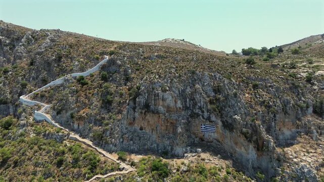 Cinematic drone approach to the Greek flag painted on the rocks of Kastellorizo. The shot follows the steep white path to the summit, revealing the mountain view. Great for travel and history