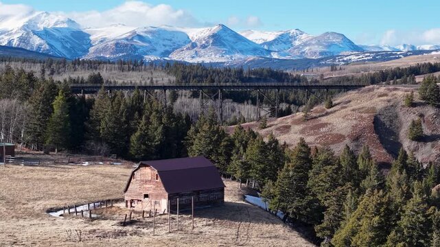 Drone footage shows a rustic barn standing on a grassy field in a rural valley with snow-capped mountains and coniferous trees near Whitefish, Montana, on a clear day.