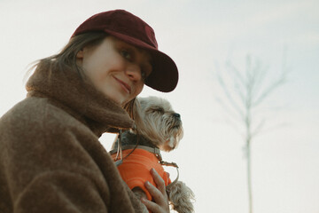 Candid portrait of a woman in a maroon cap and brown fleece jacket holding a small dog in an orange sweater. Lifestyle pet photography, emotional bond, outdoor autumn vibe, and soft natural light. © vita