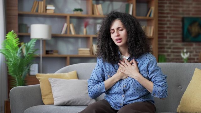 Unhealthy woman has heart attack while sitting on sofa in living room at home. Sick adult female suffers from tension in chest and massages sore spot with her hand. She needs urgent medical attention