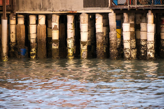 Weathered Stilts of a Traditional Stilt House Over Rippling Water. Penang Jetty construction, rough texture of barnacles on aged pillars