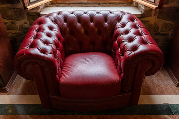 Fototapeta premium A vintage red leather tufted armchair in Chesterfield style sits beneath an open window against a stone wall, evoking a cozy, classic and nostalgic atmosphere in a warm rustic interior setting