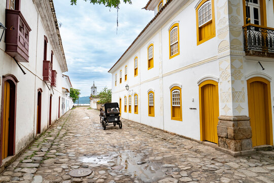 An empty historic cobblestone street featuring white colonial buildings with bright yellow windows and a horse-drawn carriage in Paraty, Brazil.