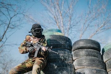Fototapeta premium Outdoor paintball scene featuring a camo-clad player with protective mask and rifle at tire cover.