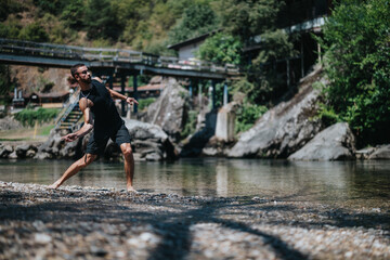 A man in a sleeveless black top and shorts balances on a rocky riverbank. The scene captures a sunny summer river setting with trees, a bridge, and calm water in the background.
