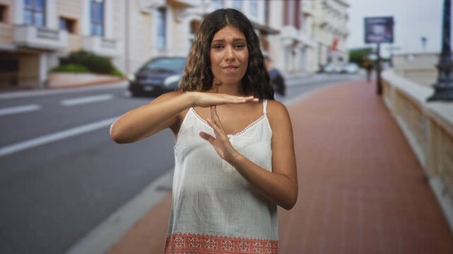 Woman in white tanktop makes time out gesture with hands on street sidewalk in front of building by parked car and lamppost; pause boundary serious.