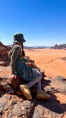 Fototapeta premium Young girl wearing a green hat and dress sits on a rock overlooking a vast desert landscape with orange sand dunes and distant mountains under a clear blue sky