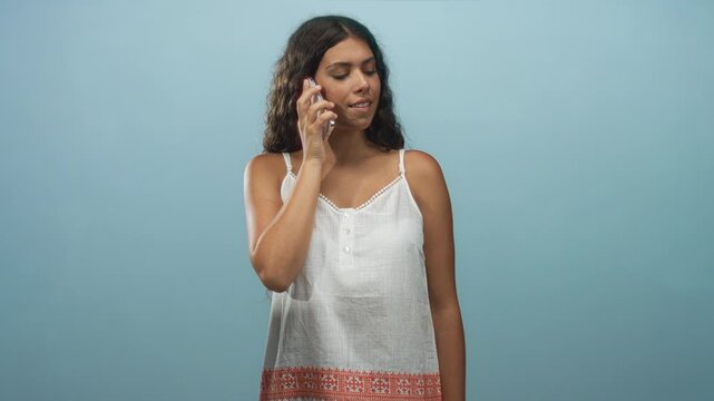 Hispanic woman holding smartphone to ear with eyes closed wearing white sundress with embroidered hem in studio; calm reflection.