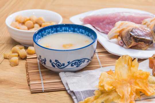 A ceramic bowl of white soup placed on a wooden table surrounded by dried scallops, pork, and mushrooms.
